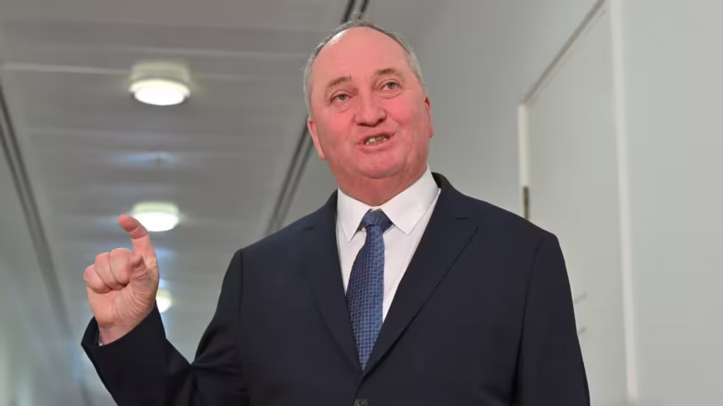 Barnaby Joyce in a blue suit and blue tie speaking with his hand raised in a white corridor.