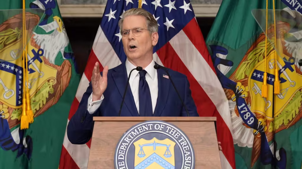 An bespectacled older man dressed in a blue suit and tie. He has greying hair and stands behind a podium with a blue and gold seal.