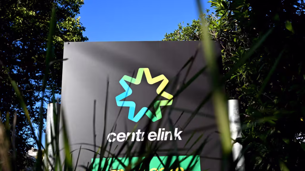 A Centrelink sign outside a building, partially obscured by grass and surrounded by trees under a clear blue sky.