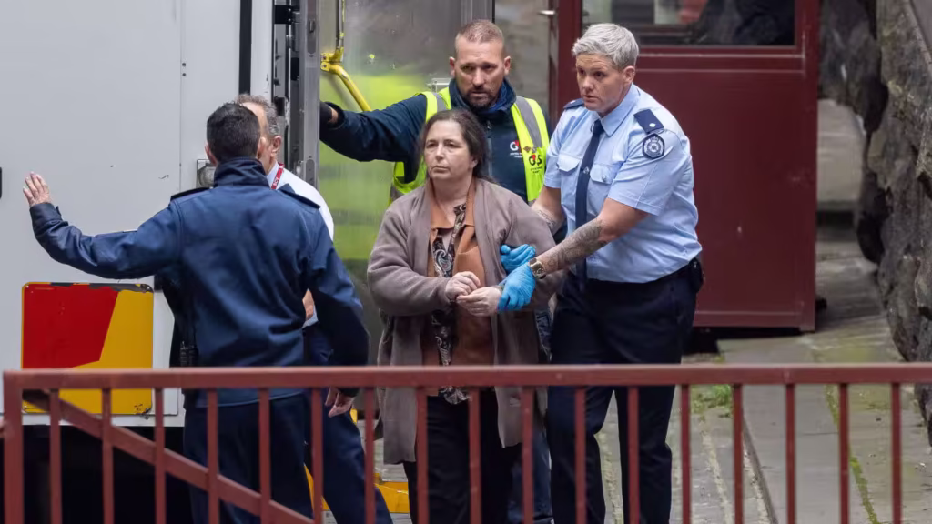 A middle-aged white woman wearing a brown cardigan is escorted off a a prison truck by blue-uniformed officers and into a courthouse.